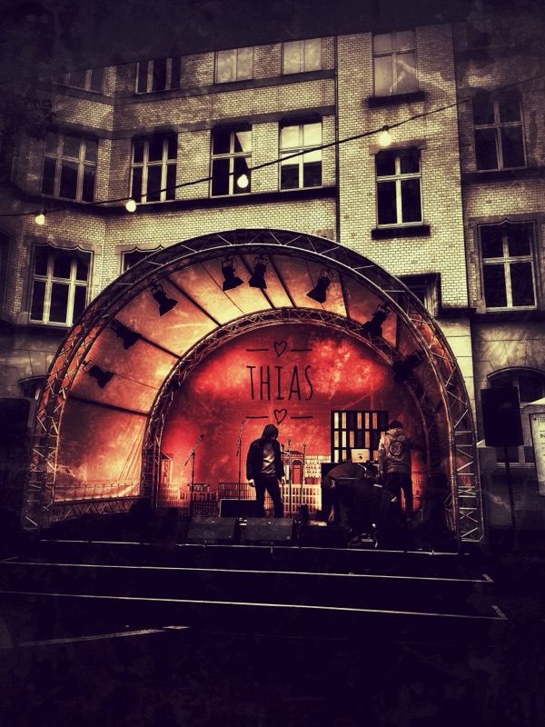 Open-Air-Bühne mit Rundbogen vor einer Backsteinfassade, auf der Rückwand der Schriftzug Thias, Thias und ein Mitmusiker auf der Bühne.