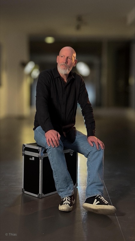 Thias sitzt in schwarzem Hemd und Jeans auf einem Flightcase in einer Halle.