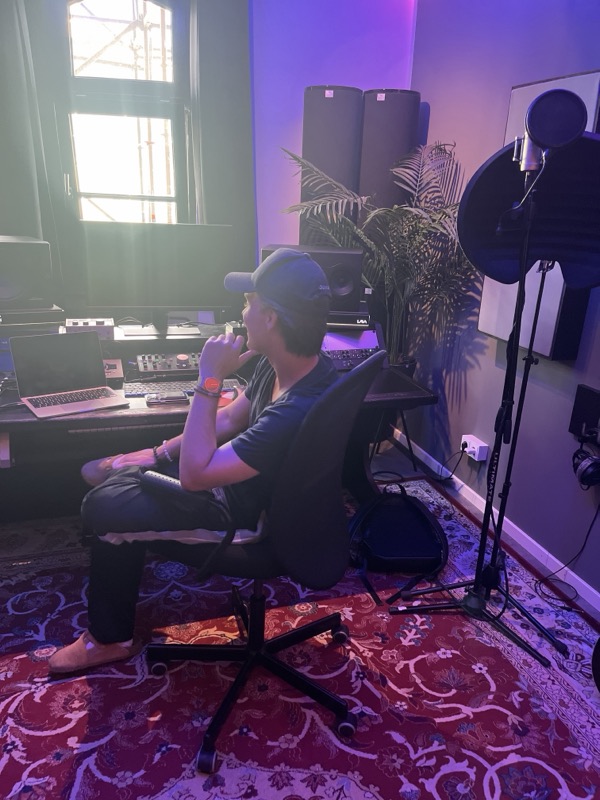 Caspar Stitz mit Kappe im Studio, er sitzt am Mischpult vor einem Fenster, daneben ein Mikrofon.