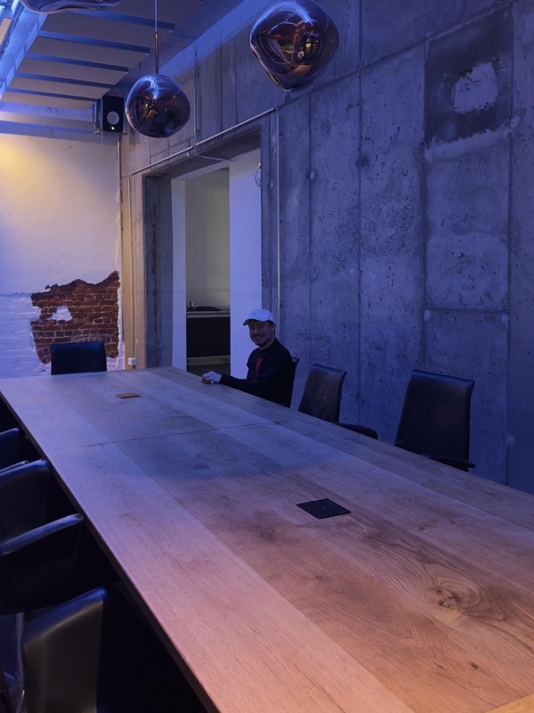 Langer Holztisch in einem Raum mit Betonwand und Kugellampen, weit hinten am Tisch sitzt Dr. Luds.
