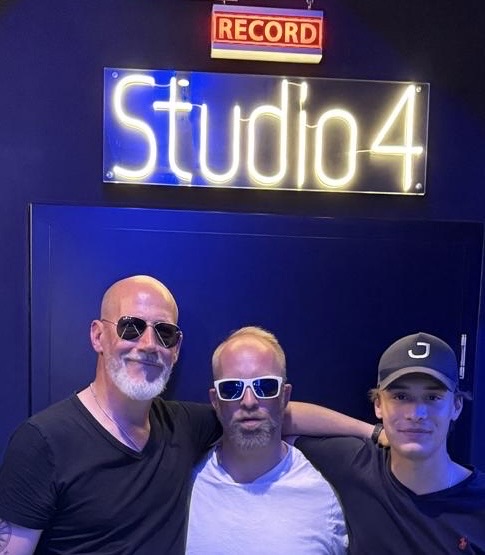 Thias, Charlante und Caspar Stitz Arm in Arm unter einem Neon-Schriftzug „Studio 4“ und einem „Record“-Schild.