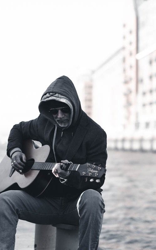 Schwarz-Weiß-Foto: Thias mit Kapuze und Sonnenbrille spielt Gitarre am Wasser.