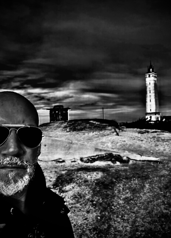 Schwarz-Weiß-Selfie: Thias mit Sonnenbrille vor einem Leuchtturm unter dunklem Himmel.