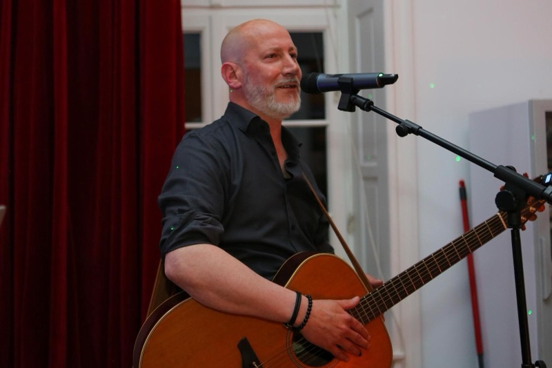 Thias live mit Akustikgitarre am Mikrofon, im Hintergrund ein roter Vorhang.