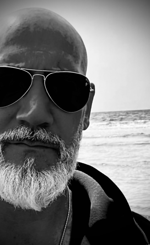 Schwarz-Weiß-Nahportrait: Thias mit Pilotenbrille und Bart, im Hintergrund das Meer.