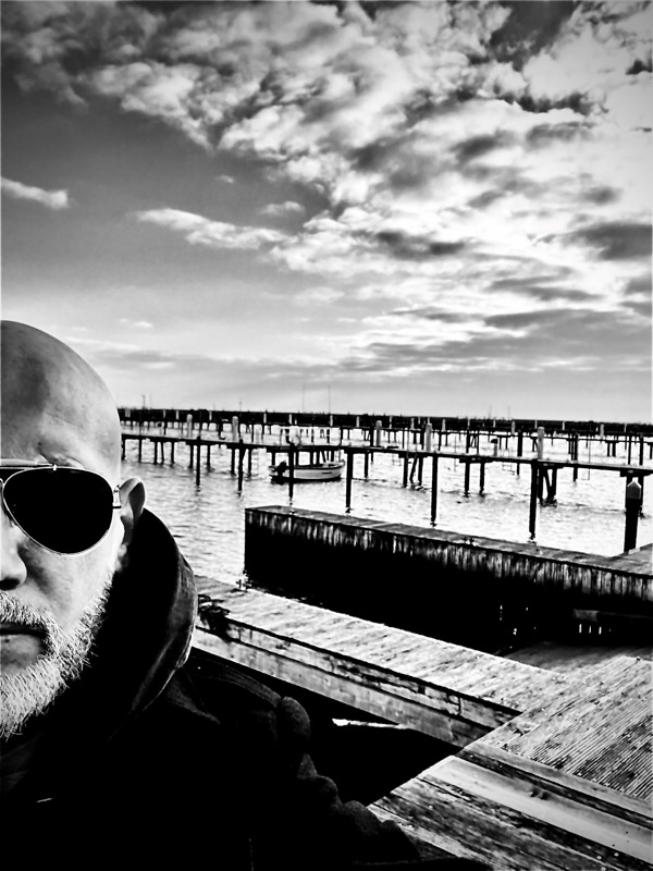 Schwarz-Weiß-Selfie: Thias mit Sonnenbrille vor einer Steganlage unter dramatischem Wolkenhimmel.