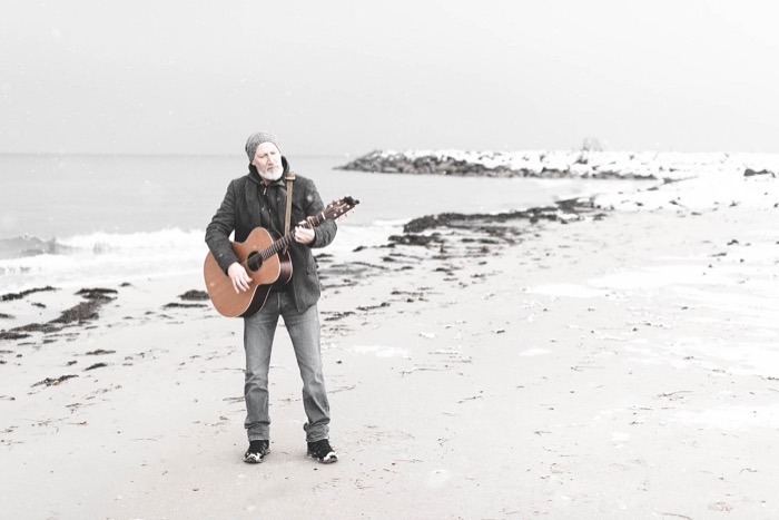 Thias spielt Gitarre an einem winterlichen, hellen Strand.