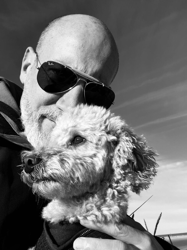 Schwarz-Weiß-Foto: Thias mit Sonnenbrille und grauem Bart hält einen lockigen, hellen Hund im Arm, dahinter der Himmel.